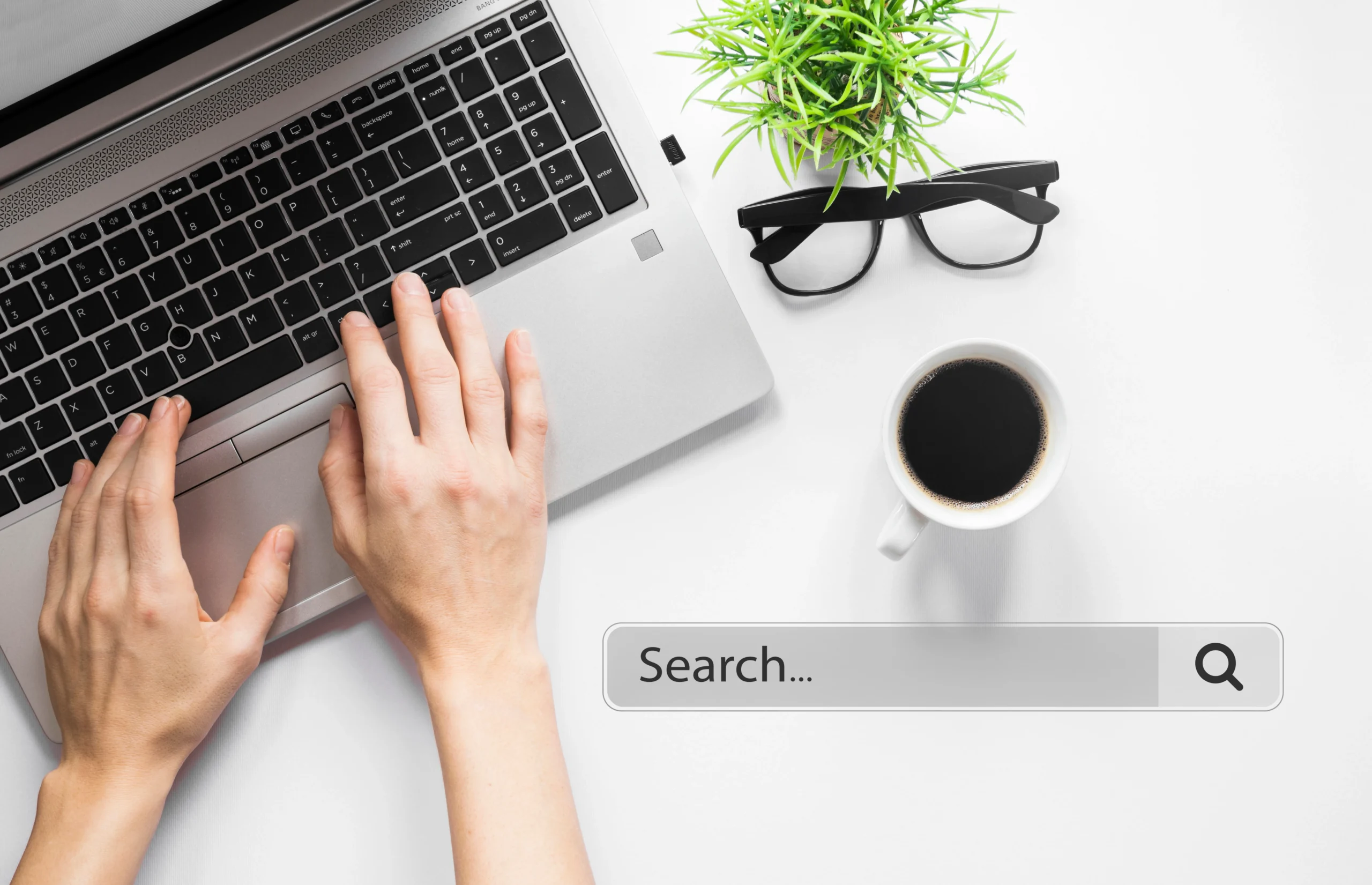 Person typing on a laptop with a search bar, coffee cup, glasses, and plant on a white desk.