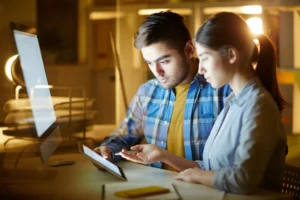 Two professionals reviewing content on a tablet while working at a computer in an office, focusing on digital strategy and collaboration.