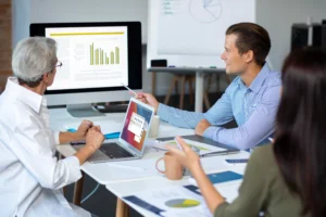 Three colleagues in a meeting reviewing charts and graphs on a computer and laptop in an office setting.